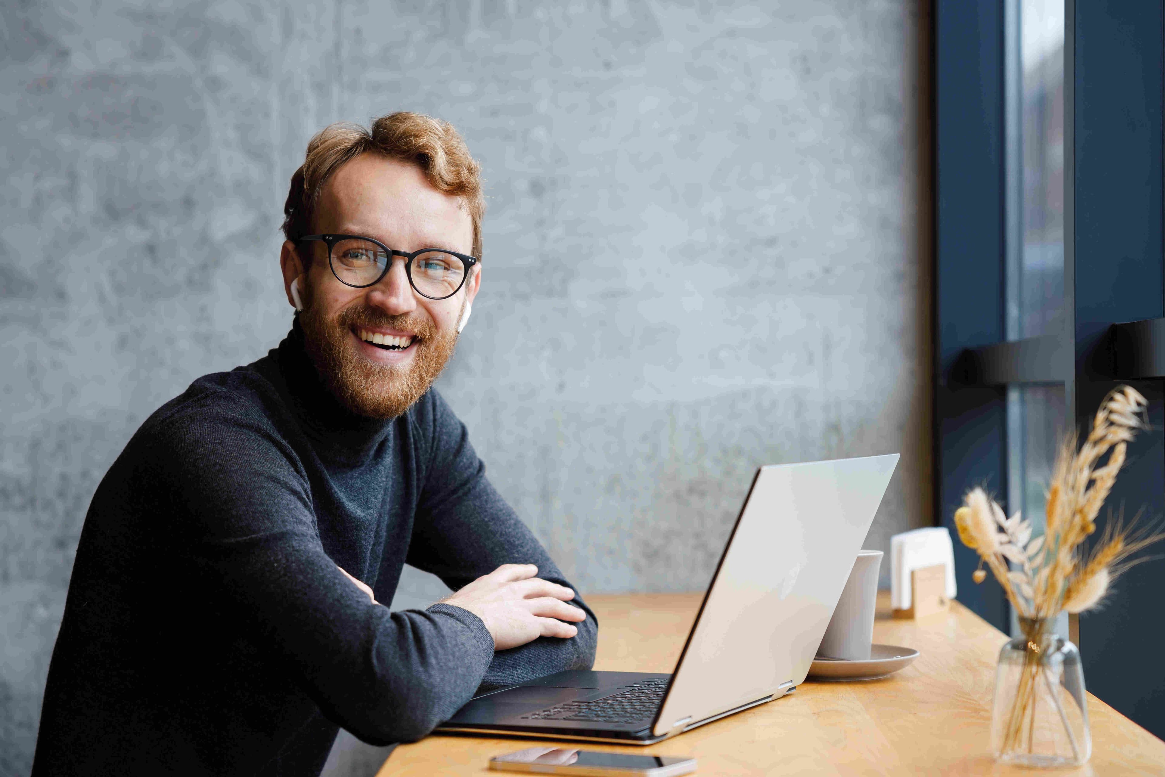 Mladý podnikatel s brýlemi pracuje na notebooku ve stylové kavárně. Usmívá se do kamery a ztělesňuje ducha freelancingu, práce na dálku a moderní online komunikace pro malé podniky.